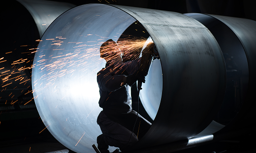 Ein Mann schweißt ein großes Metallrohr in einer Werkstatt, umgeben von Werkzeugen und Schweißgeräten.